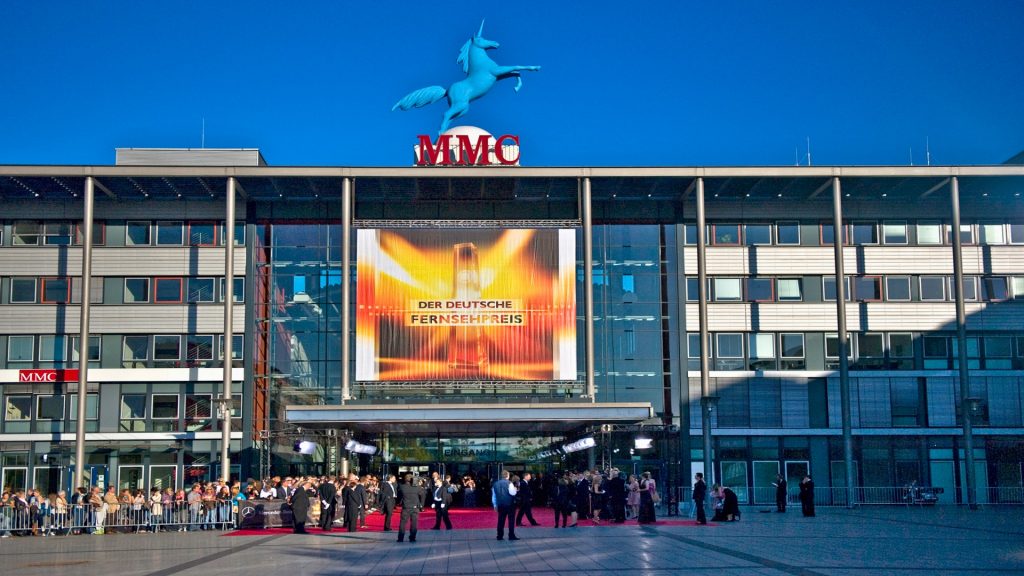 Der große Haupteingang der MMC Studios Köln mit dem Einhorn-Logo auf dem Dach und vielen Menschen die neben dem roten Teppich stehen