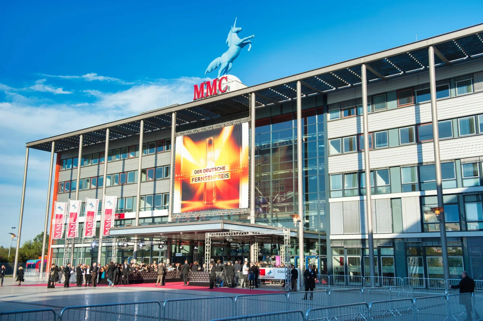 Der große Haupteingang der MMC Studios Köln mit dem Einhorn-Logo auf dem Dach und vielen Menschen die neben dem roten Teppich stehen